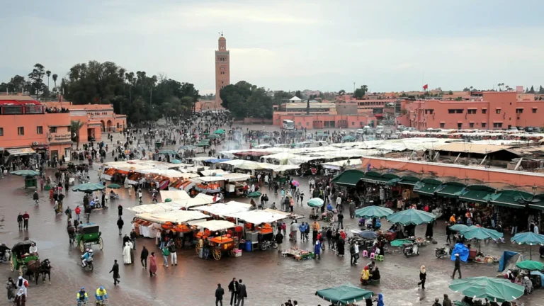 Vue d'ensemble de la place Jemaa el-Fna à Marrakech en journée, étals de marché aux auvents blancs et oranges, calèches et minaret de la mosquée Koutoubia à l'arrière-plan