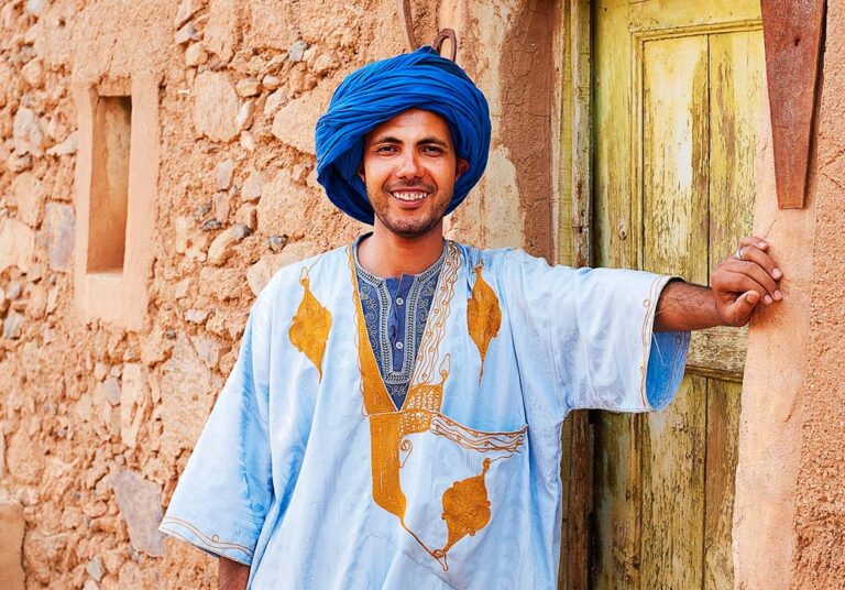 Un homme amazigh (berbère) souriant en turban bleu et gandoura traditionnelle brodée, devant une porte ancienne et un mur en pisé au Maroc