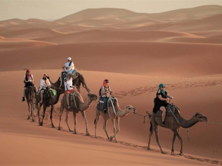 Caravane de touristes à dos de chameau, en turban coloré, traversant les dunes de l'Erg Chebbi au coucher du soleil près de Merzouga