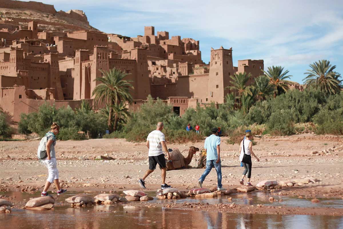 Travellers cross the Ounila River on stepping stones to reach the UNESCO ksar of Aït Ben Haddou — a Day 1 highlight of the Shared 3-Day Marrakech to Merzouga Desert Tour.