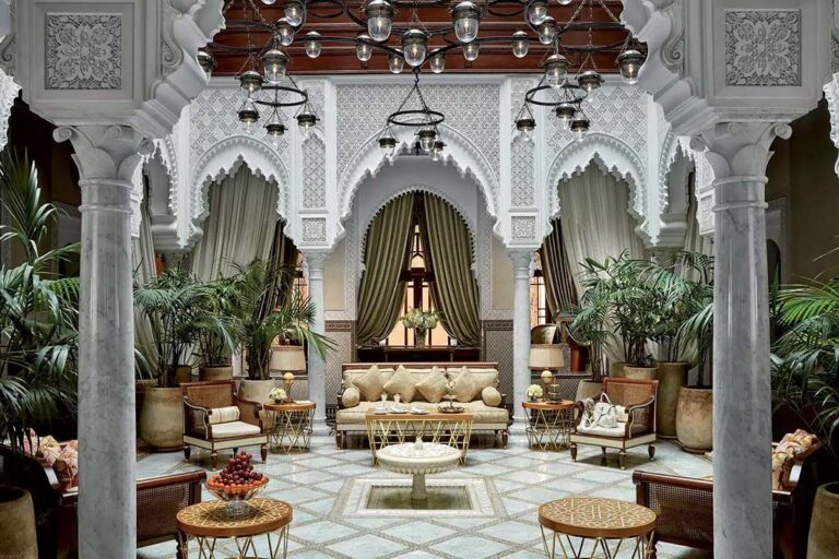 Patio central d'un riad de luxe à Marrakech : arcades en stuc ciselé, colonnes de marbre, zellige au sol et fontaine centrale entourée de palmiers